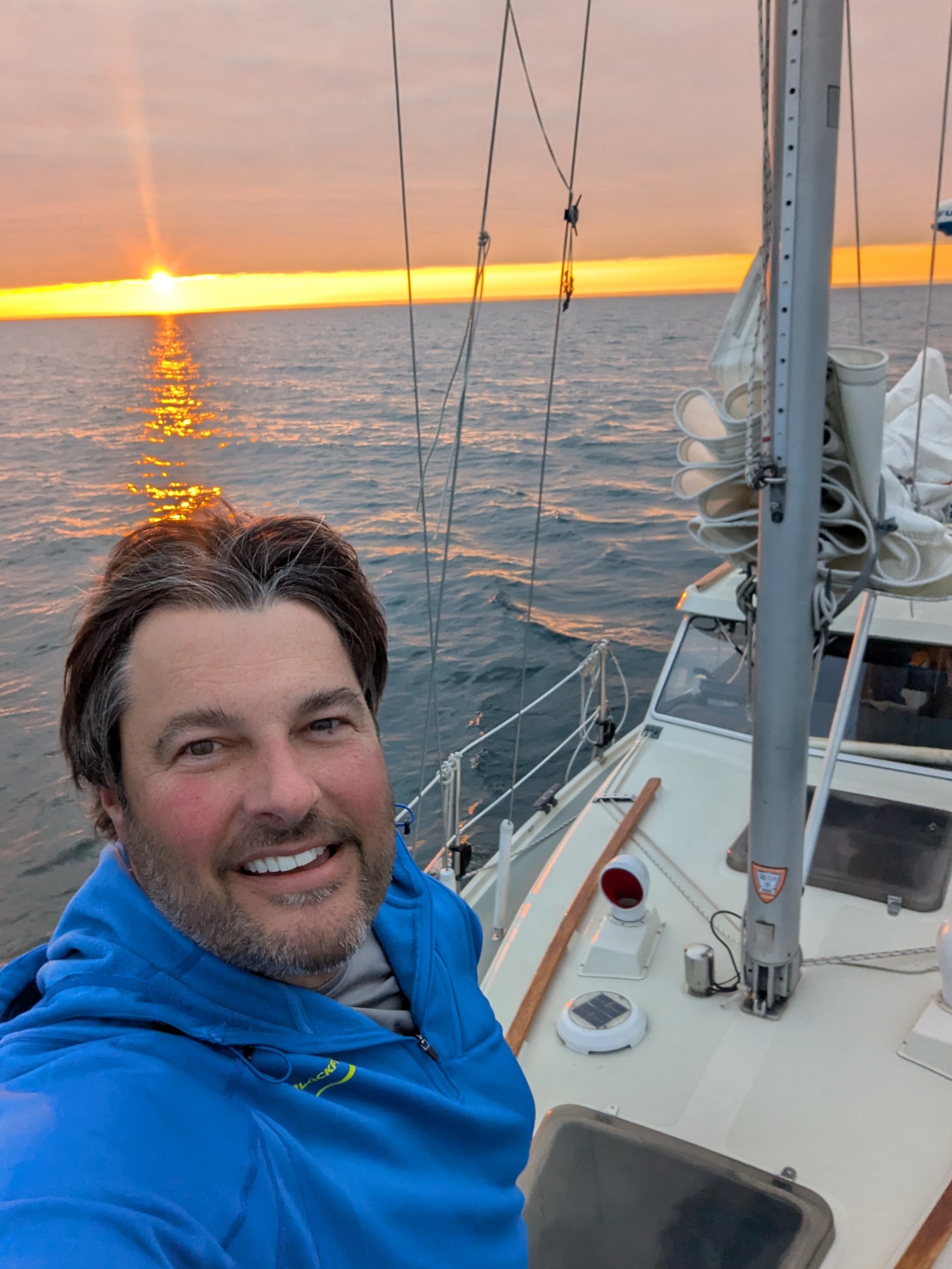 Smiling man in blue jacket taking a selfie on a boat at sunset.