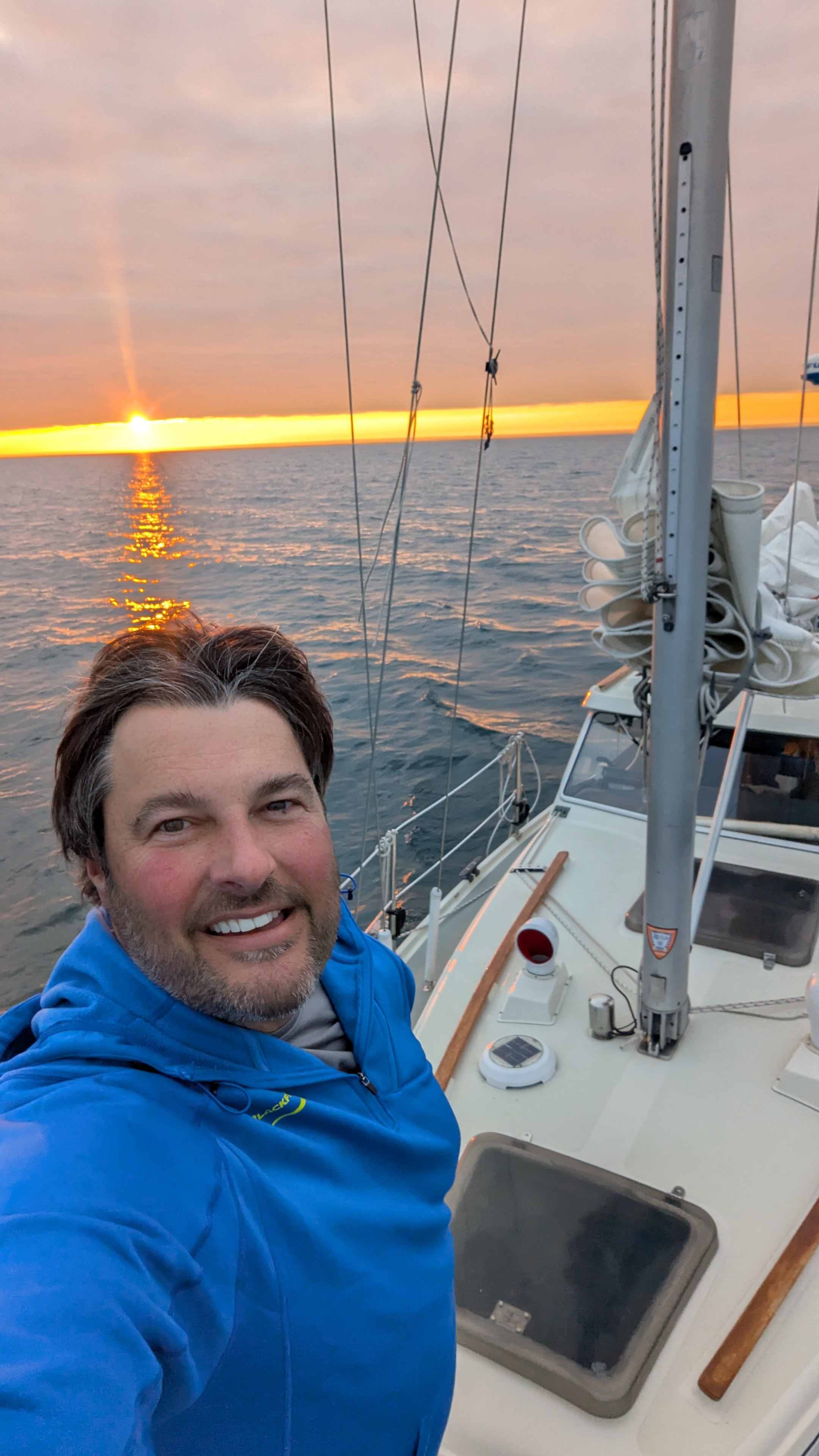Smiling man in blue jacket taking a selfie on a boat at sunset.
