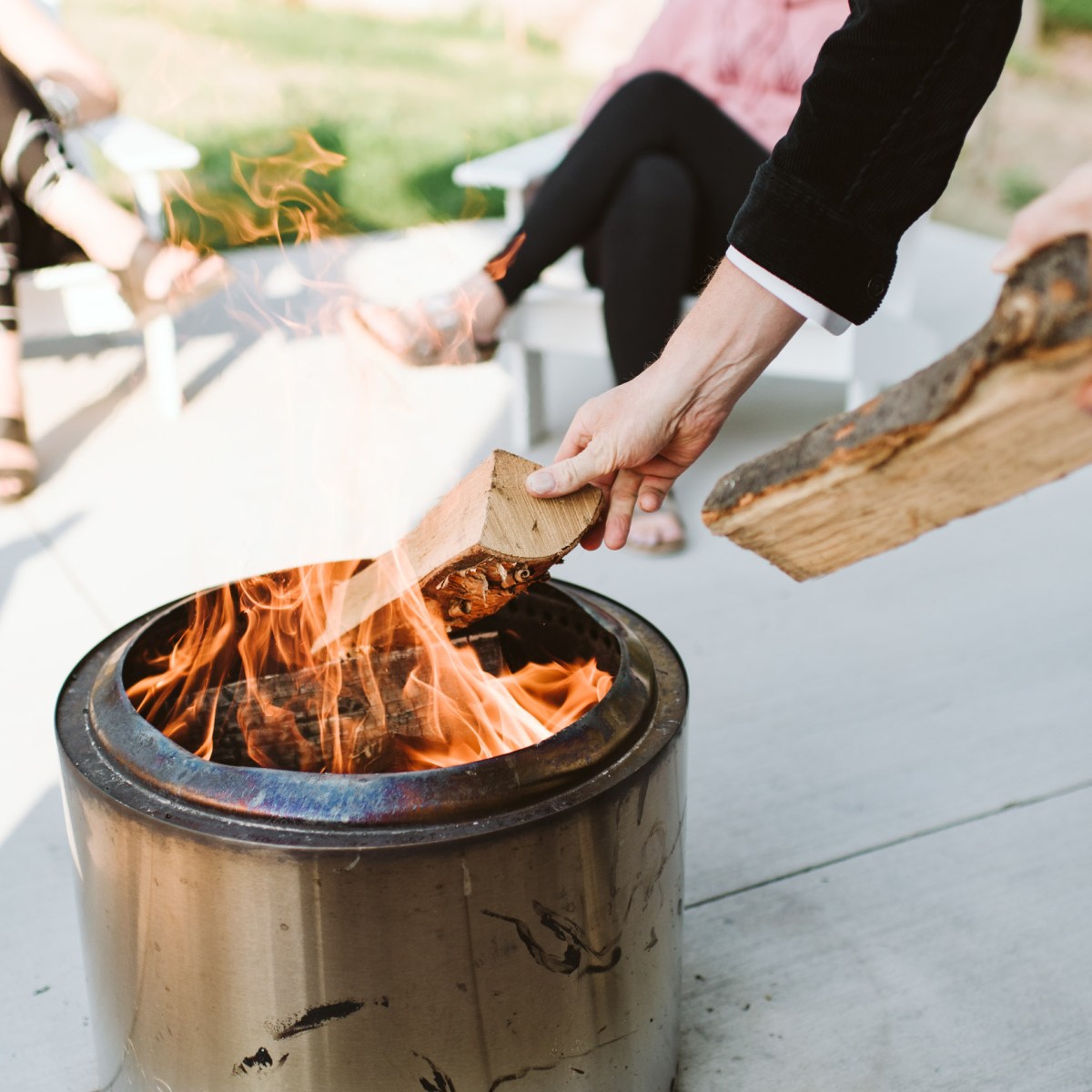 Person adding wood to a metal fire pit, seated people in background.