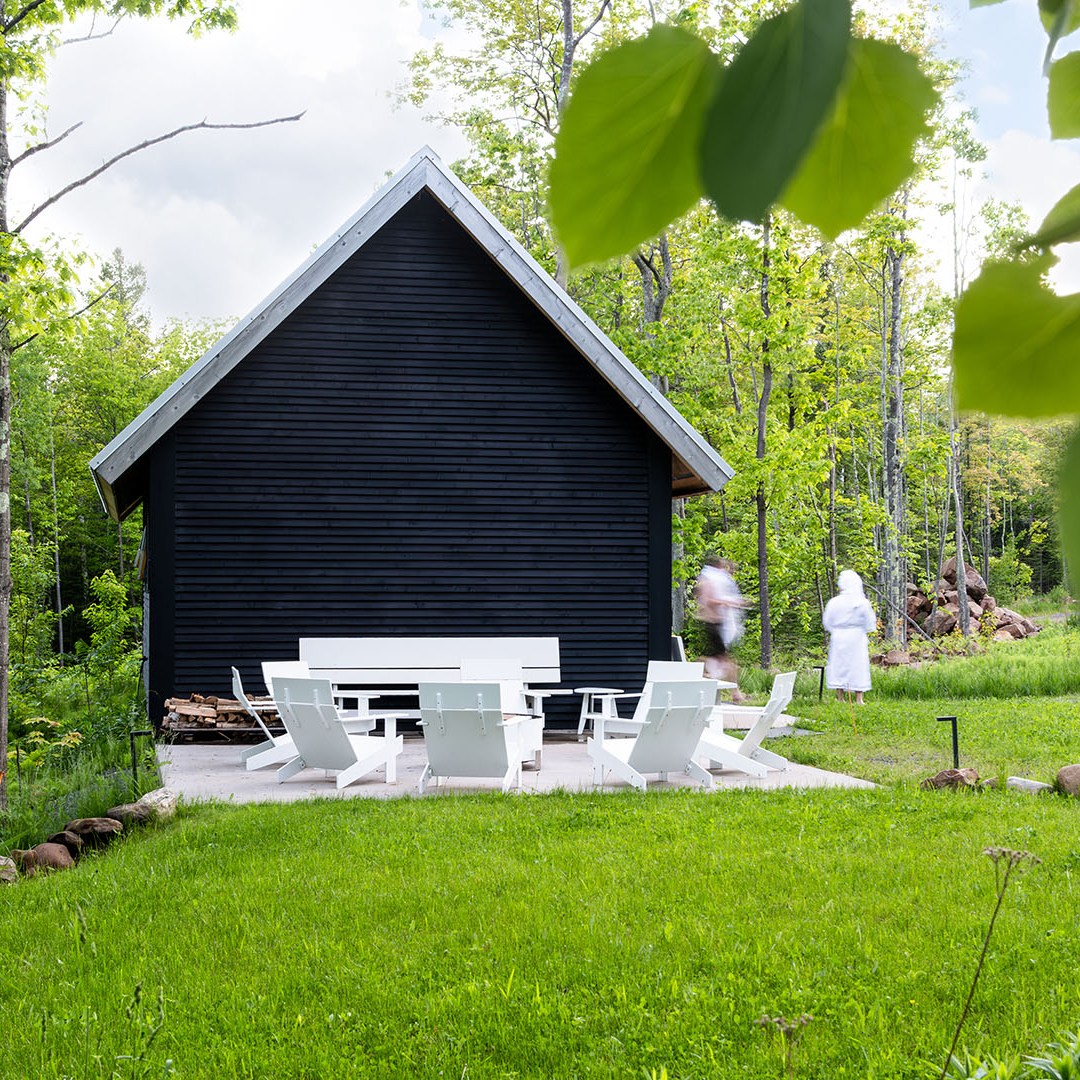 Modern black cabin with white patio chairs, set in a lush green forest.