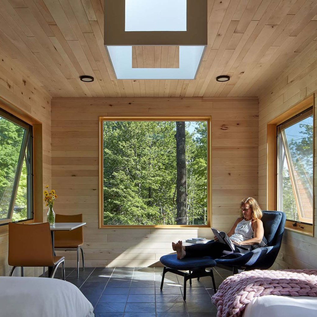 Woman relaxing with a tablet in a wooden cabin, large windows showing trees, skylight above.