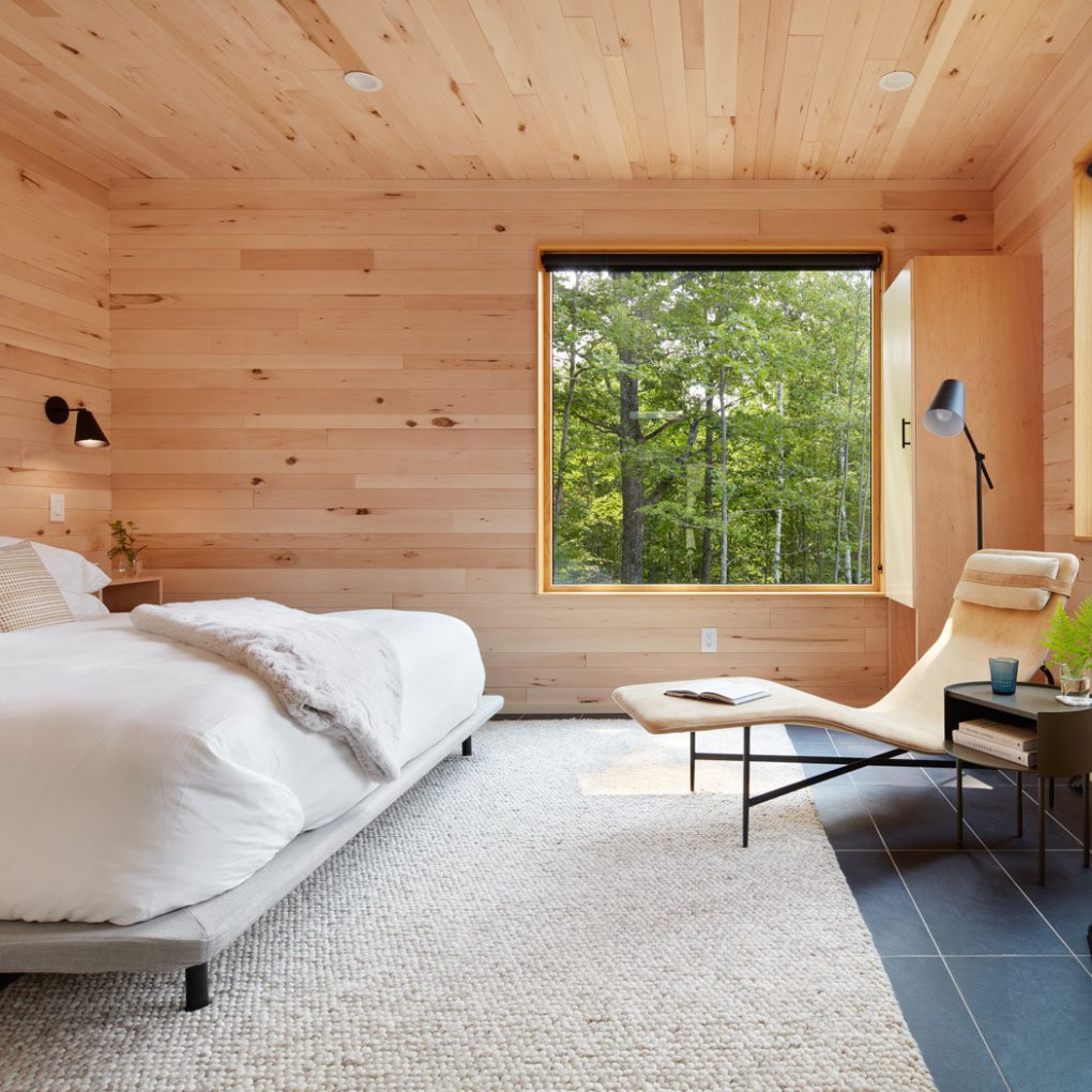 Modern bedroom with wood walls, large windows, white bed, lounge chair, and small side table.
