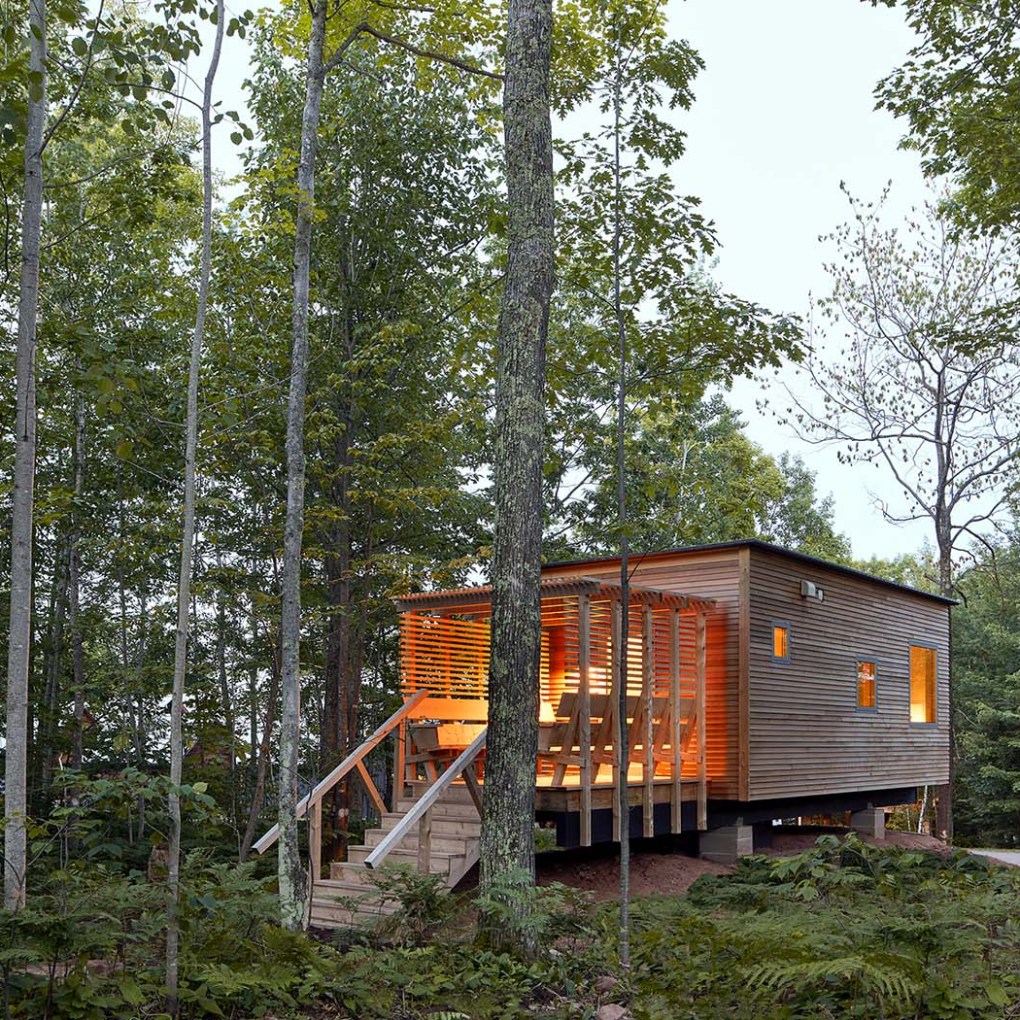 Small cabin with porch in a forest setting, surrounded by tall trees.